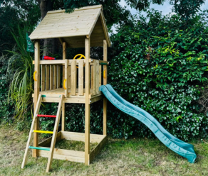 Compact houten speelhuisje met groene glijbaan en klimladder naast een heg